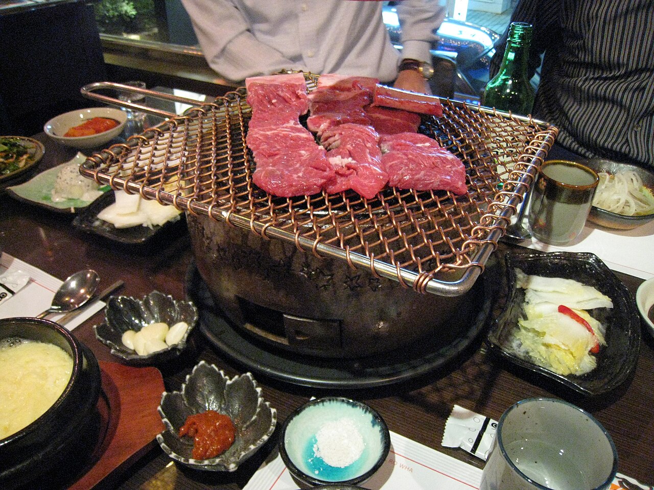 Raw galbi on a charcoal table grill, surrounded by side dishes