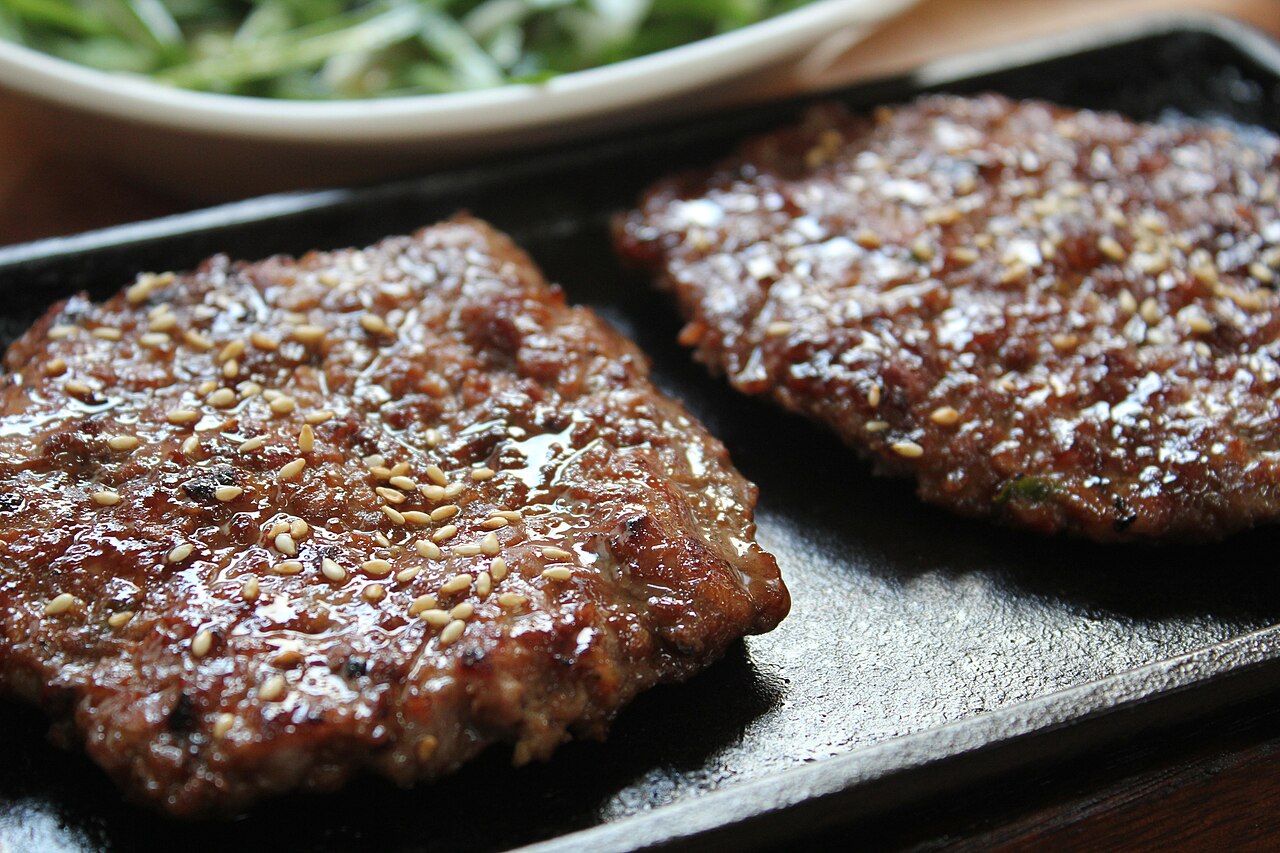 Two grilled tteok-galbi patties with a seeded glaze on a hot plate