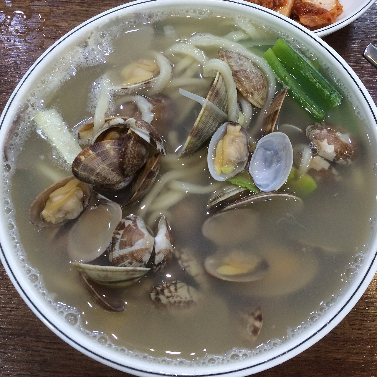 A bowl of clam kalguksu with many clams in their shells, noodles and scallion in a clear broth