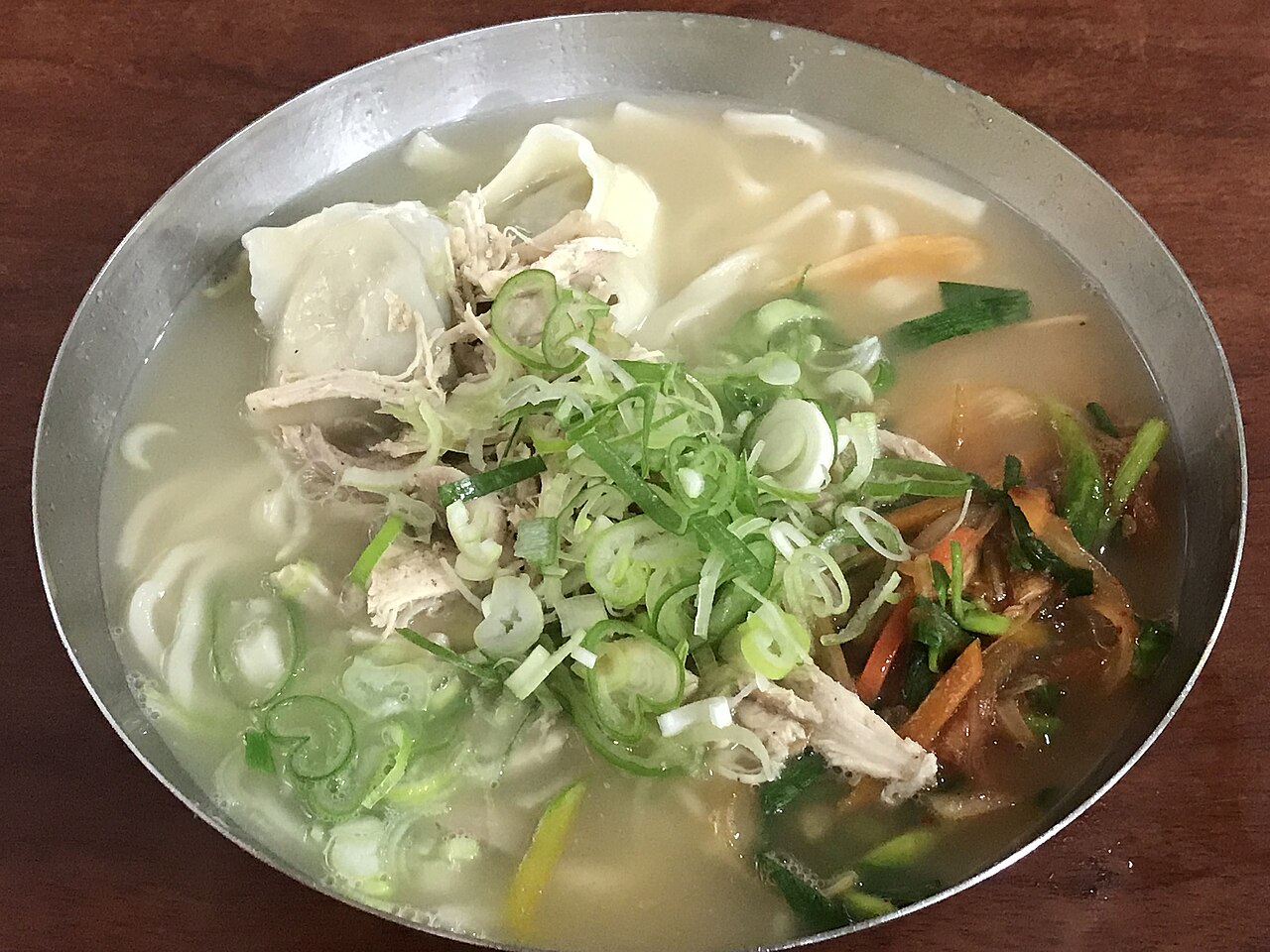 A metal bowl of chicken kalguksu with shredded chicken, dumplings, wide noodles and chopped scallions
