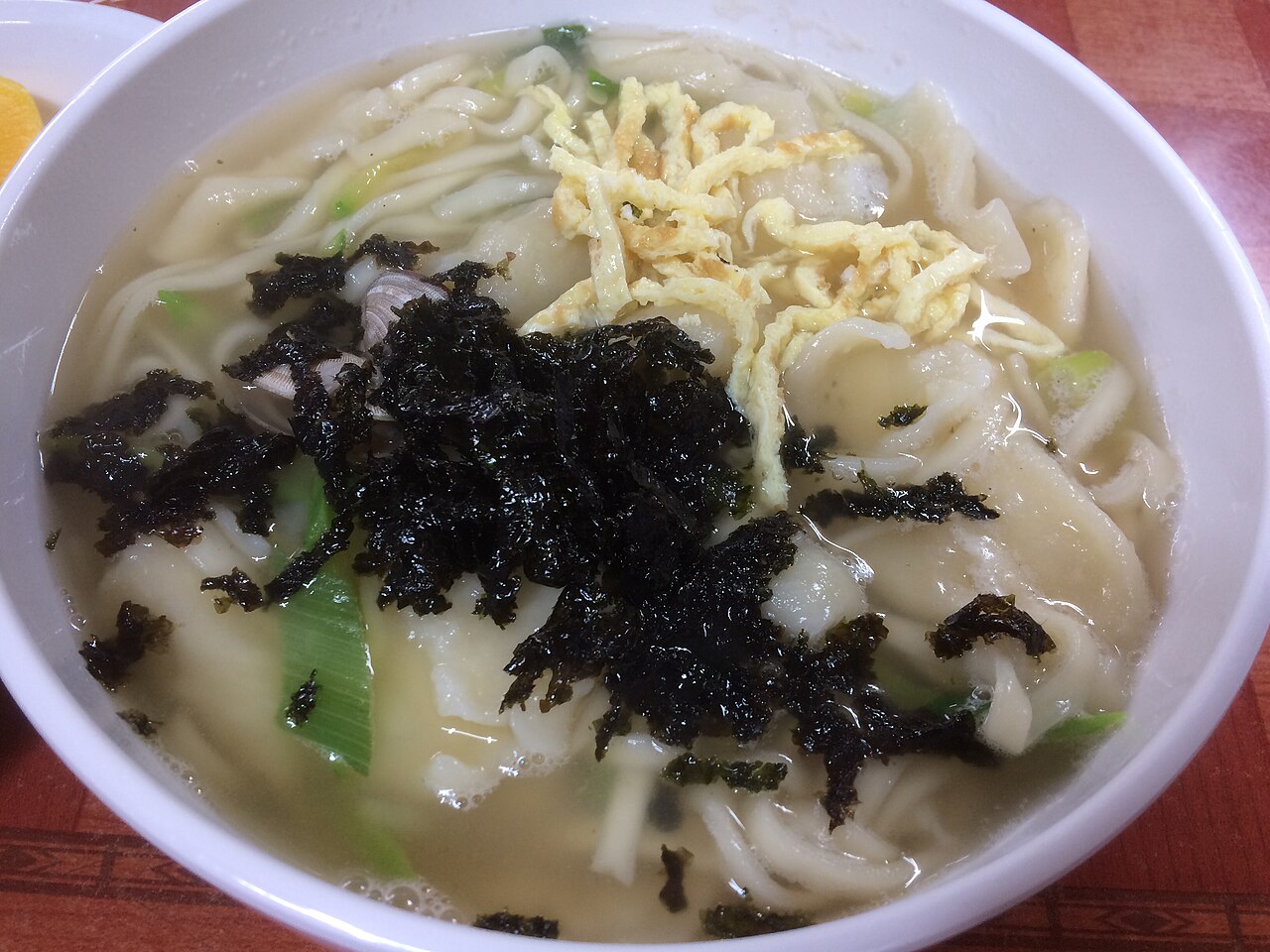 A bowl of kaljebi with knife-cut noodles and torn dough flakes, egg strips, seaweed and a clam in the broth
