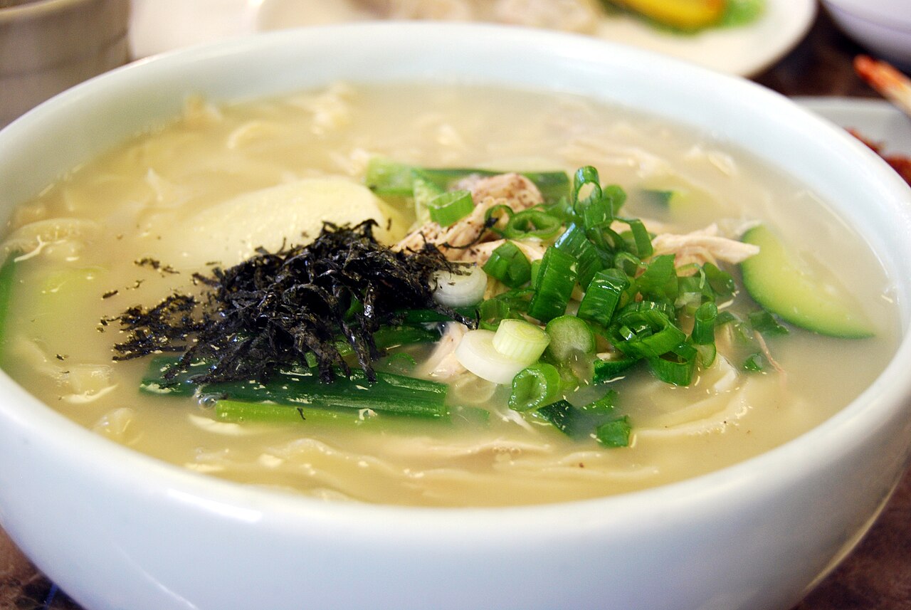 A bowl of kalguksu in a clear broth with chicken, zucchini, scallions and shredded seaweed