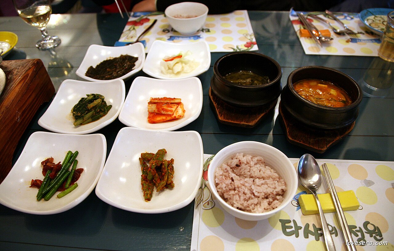 A baekban table with rice, a soup, a red stew in a stone pot and several small dishes of vegetables and kimchi