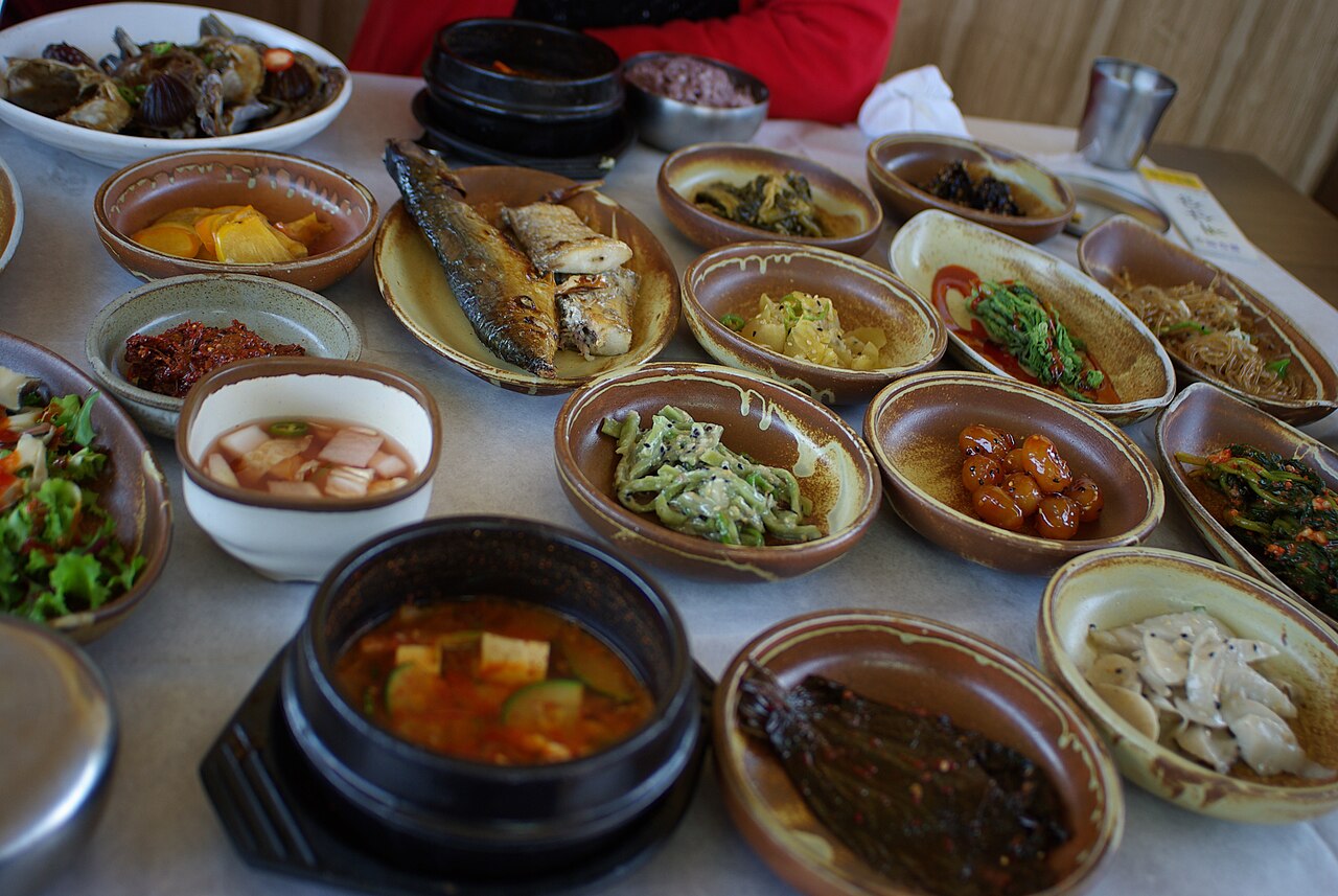 A table full of small bowls of side dishes, grilled fish and a red stew