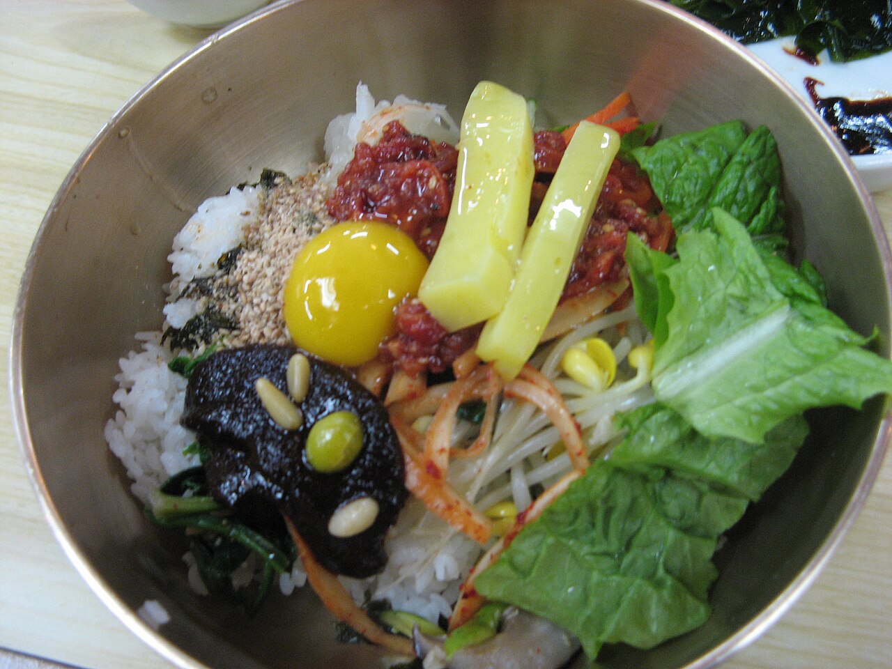 A metal bowl of bibimbap with rice, an egg yolk, chili paste, bean sprouts, lettuce and pickled radish