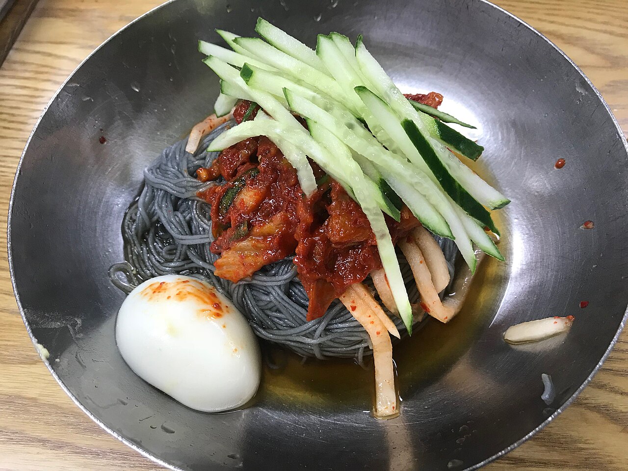 Bibim-naengmyeon: dark noodles topped with spicy sauce, cucumber and a boiled egg in a steel bowl