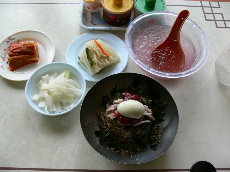 Bibim makguksu in a dark bowl with side dishes of kimchi and pickled radish