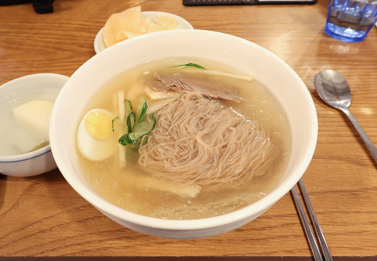 Pyeongyang-style naengmyeon in a white bowl, with pale buckwheat noodles in clear broth