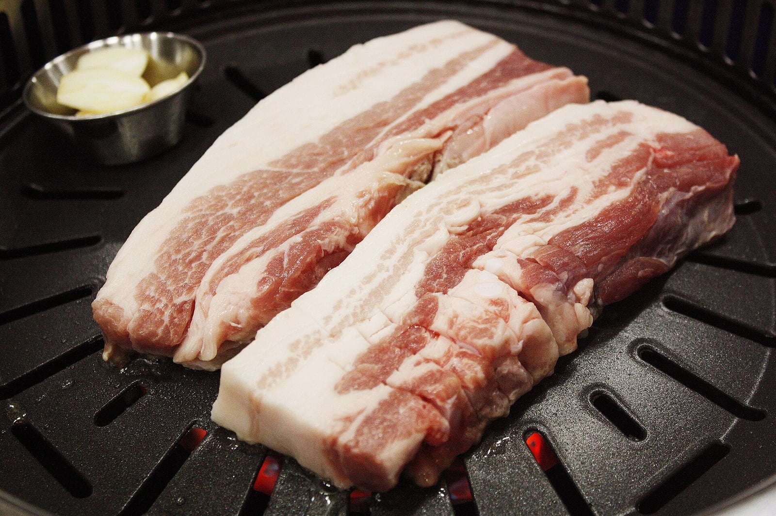 Two thick slices of raw pork belly on a round grill plate with sliced garlic