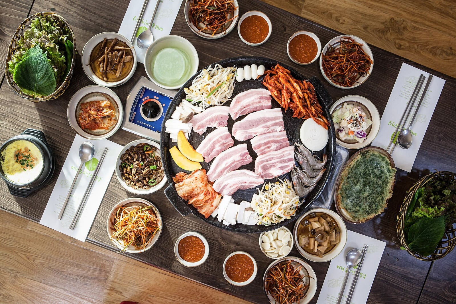 Raw samgyeopsal pork belly on a table grill, surrounded by side dishes, lettuce and sauces