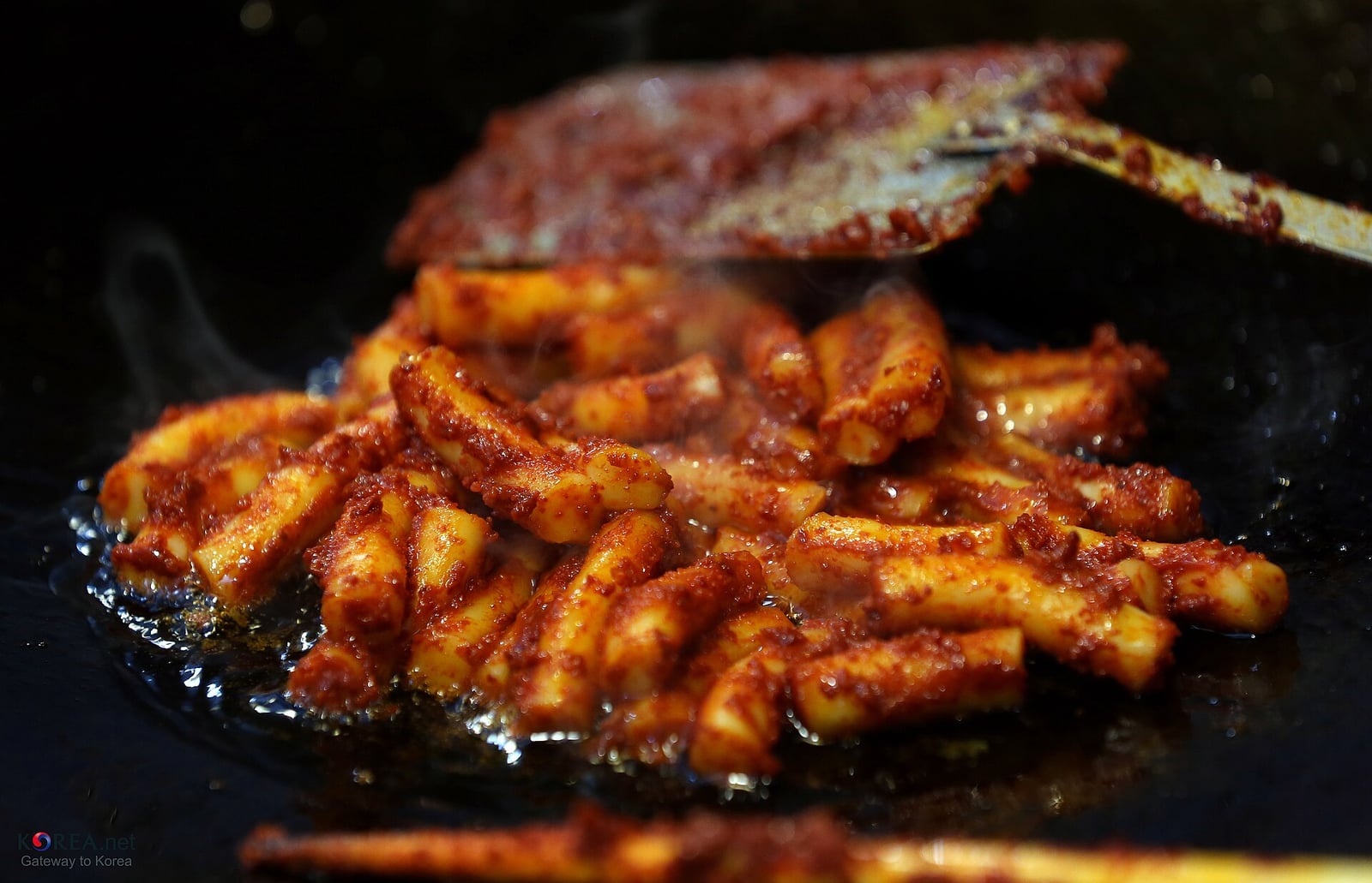 Rice cakes frying in red seasoning on a hot griddle with a spatula
