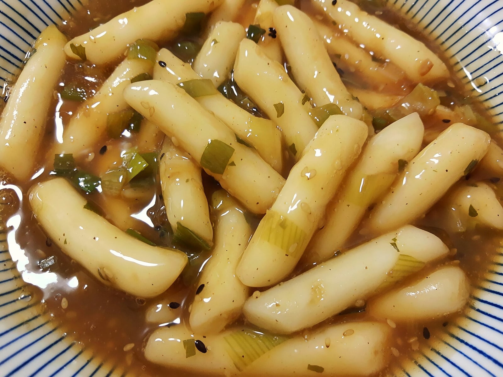 Soy sauce tteokbokki with sliced scallion and sesame seeds in a striped bowl